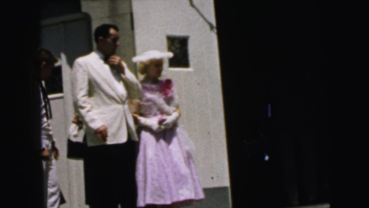 In 1964 Camden, New Jersey footage, a groom in a white dinner jacket pauses beside a woman in a lavender dress and white hat outside a church.