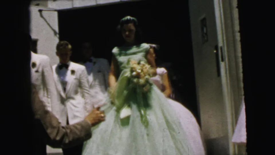 A bride in a pale green gown and veil steps forward with a bouquet while attendants in white jackets and pastel dresses remain near the Camden church doorway.