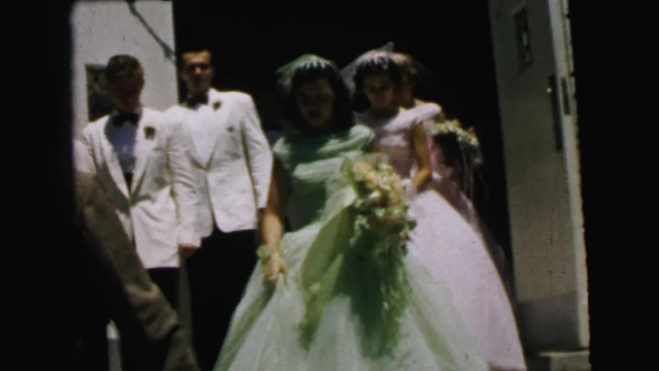 The bride in a pale green gown stands with bouquet near groomsmen in white jackets and another woman in pink outside the Camden church entrance.