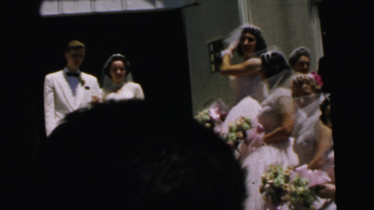 Bride and groom in formal wedding attire stand near a Camden church doorway as attendants and guests partially block the home-movie view.