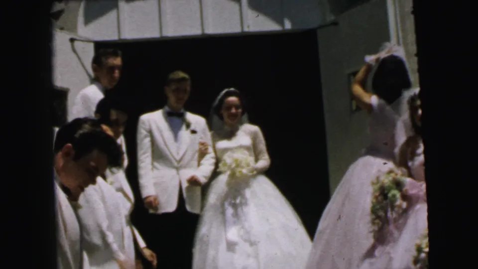 Bride and groom stand arm in arm outside the church in 1964 Camden, framed by attendants, veils, bouquets, and a dark doorway.