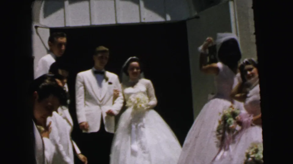 In Camden, New Jersey wedding footage from 1964, the bride and groom stand arm-in-arm near a church doorway as attendants gather around them.