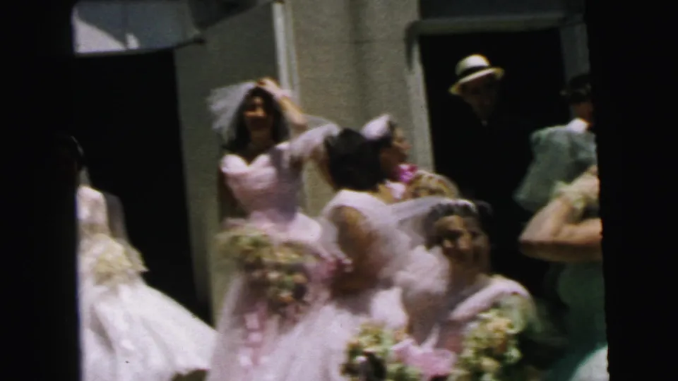 1964 Camden home-movie frame showing a bride, groom, and pink-dressed bridesmaids clustered outside a doorway with bouquets.