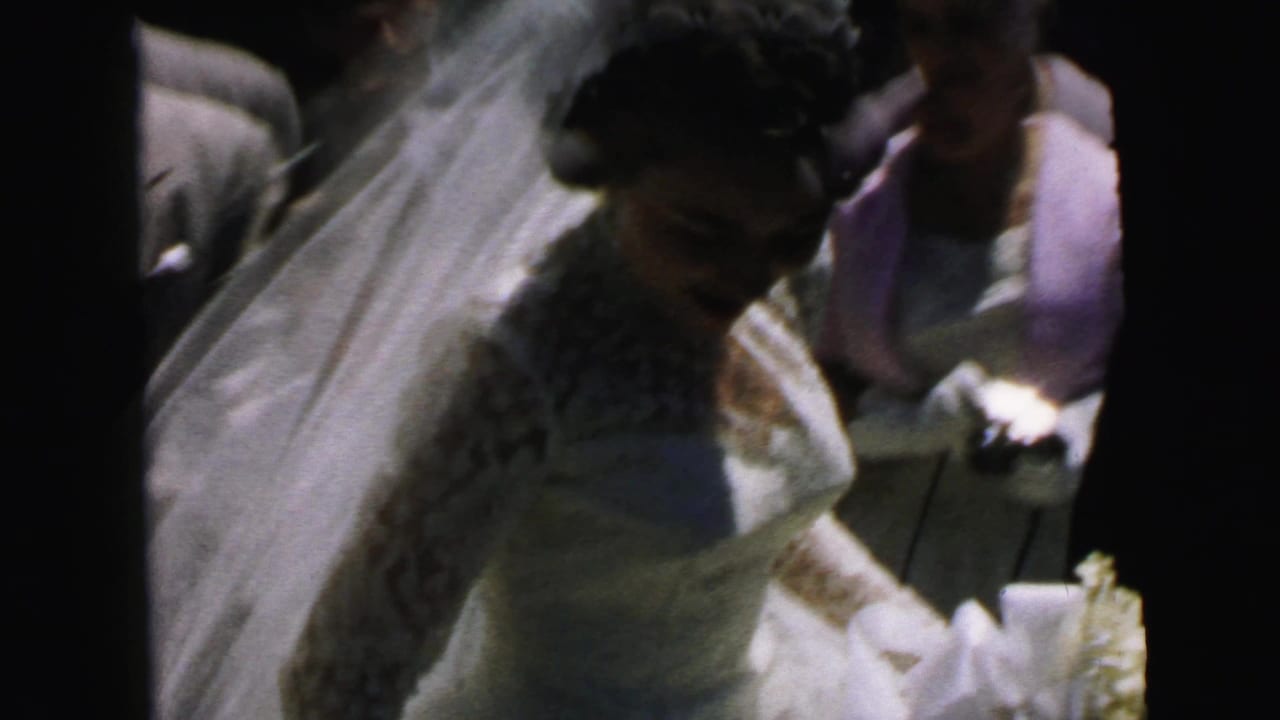Bride in a white dress and veil stands beside guests near a car.