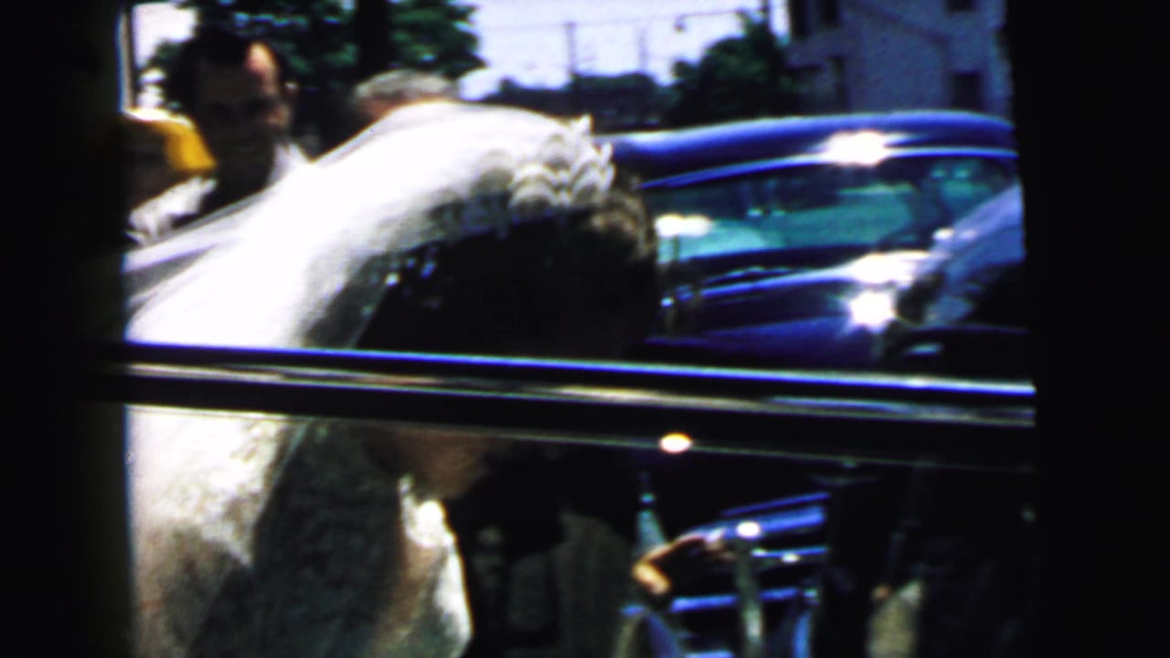 Bride bends toward the open door of a dark car as guests stand nearby.