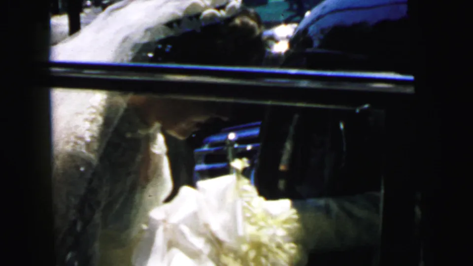 Bride in white dress and veil is partly obscured by the dark sedan door as she gets in.