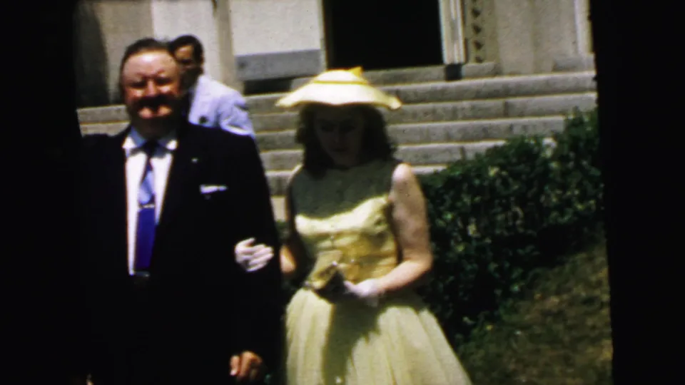 Wedding guests, including a suited man and a woman in a pale dress and hat, stand outside near steps in Camden.