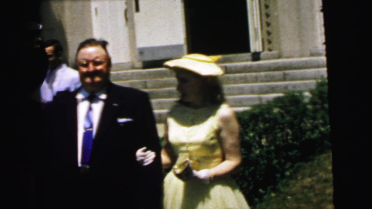 In 1964 Camden footage, a man in a dark suit poses beside a woman in a yellow dress and matching hat outside stone steps.