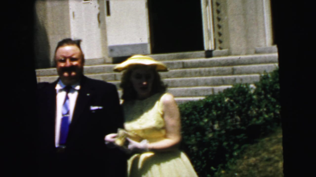 The woman in a yellow dress and hat stands arm-in-arm with the suited man near stone steps in the Camden home-movie scene.