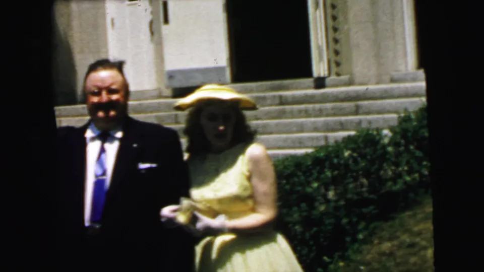 The woman in a yellow dress and hat stands arm-in-arm with the suited man near stone steps in the Camden home-movie scene.