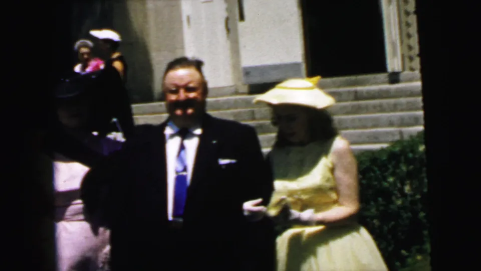 A well-dressed person at the edge of frame joins the view as the Camden 1964 couple remains posed outside the stone building.