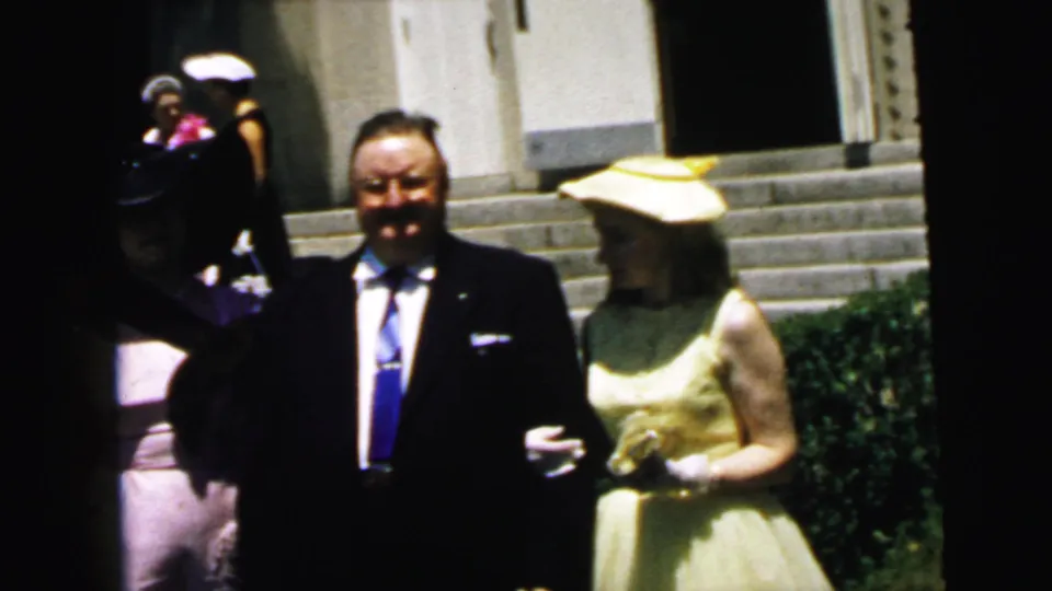 The suited man and woman in yellow remain outside the stone building while another formally dressed guest moves through the left side of frame.