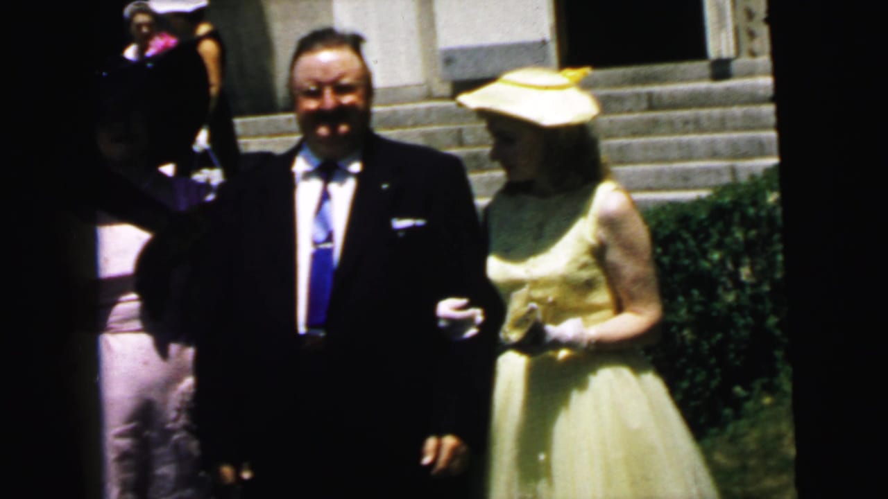 1964 Camden home-movie frame showing adults in formal wear near church steps, with a dark-suited man and a woman in a yellow dress and hat.