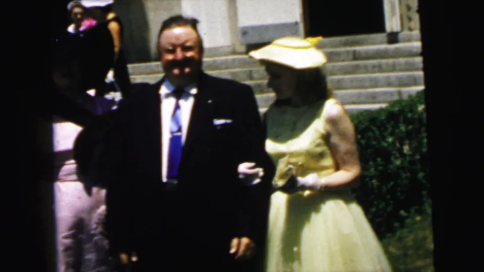 1964 Camden home-movie frame showing adults in formal wear near church steps, with a dark-suited man and a woman in a yellow dress and hat.