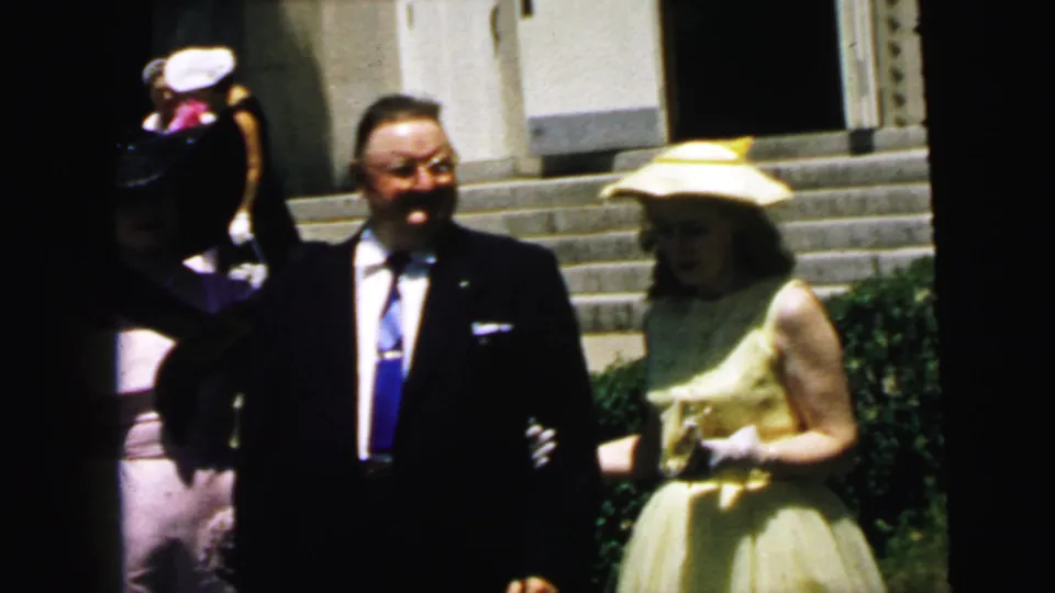 1964 Camden home-movie frame of a dark-suited man and a woman in a yellow dress waiting near stone church steps and greenery.