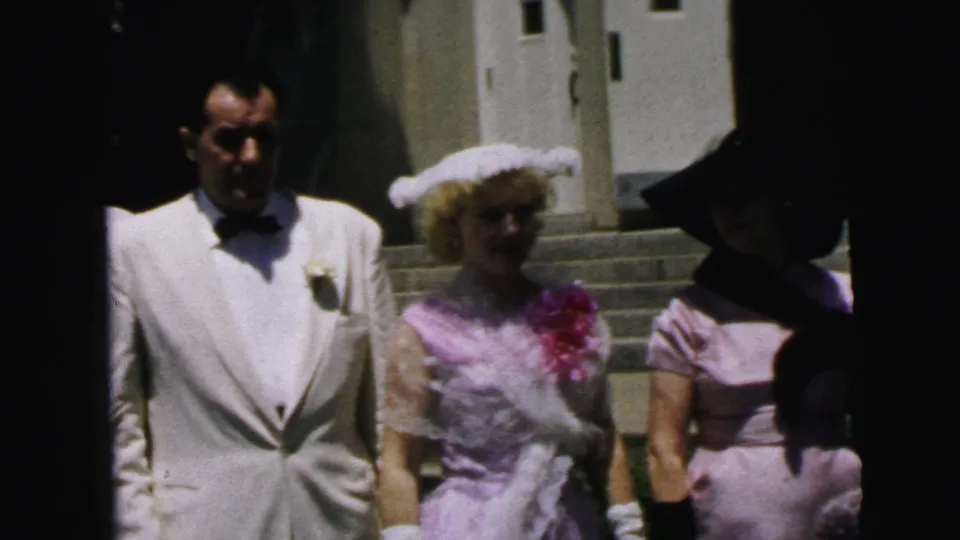 1964 Camden church exterior frame showing a man in a light tuxedo with women in pink and pale dresses near the entrance.