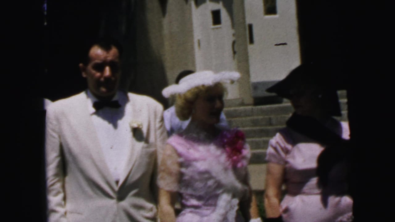 In Camden, New Jersey, 1964 footage, a man in a white tuxedo walks beside a woman in a pink dress and white hat outside a building.