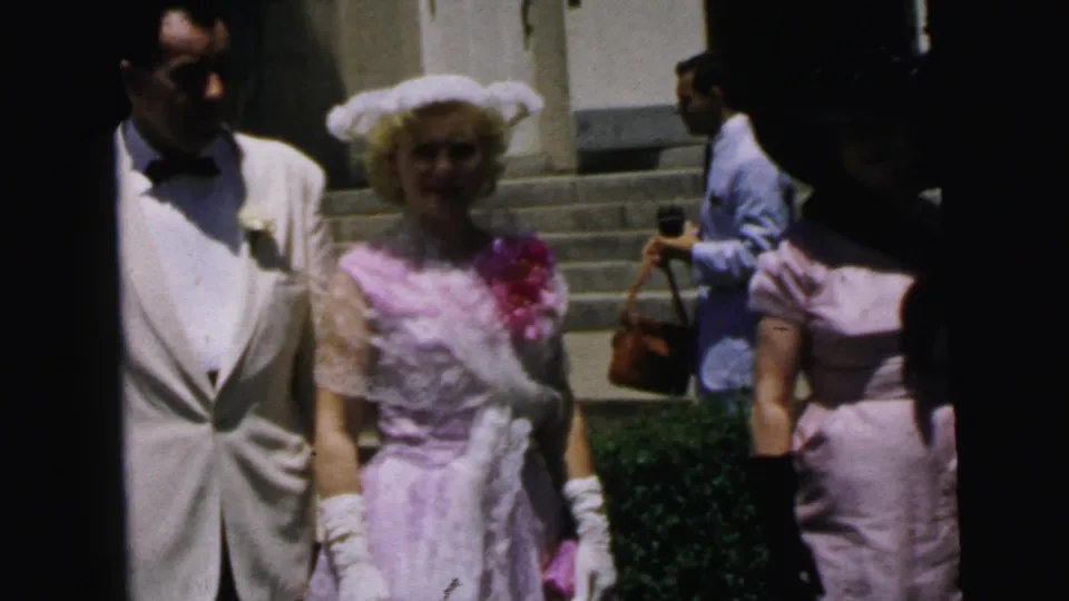 A white-jacketed man and a woman in a pink formal dress face the camera near Camden building steps, with another adult visible in the background.
