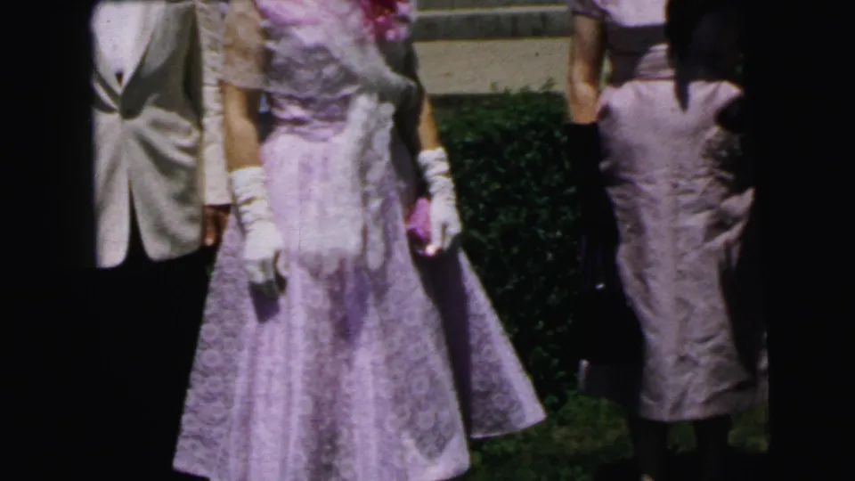 In the closing frame, the pink-dressed woman with white gloves moves near a companion in a dark hat outside the Camden building.