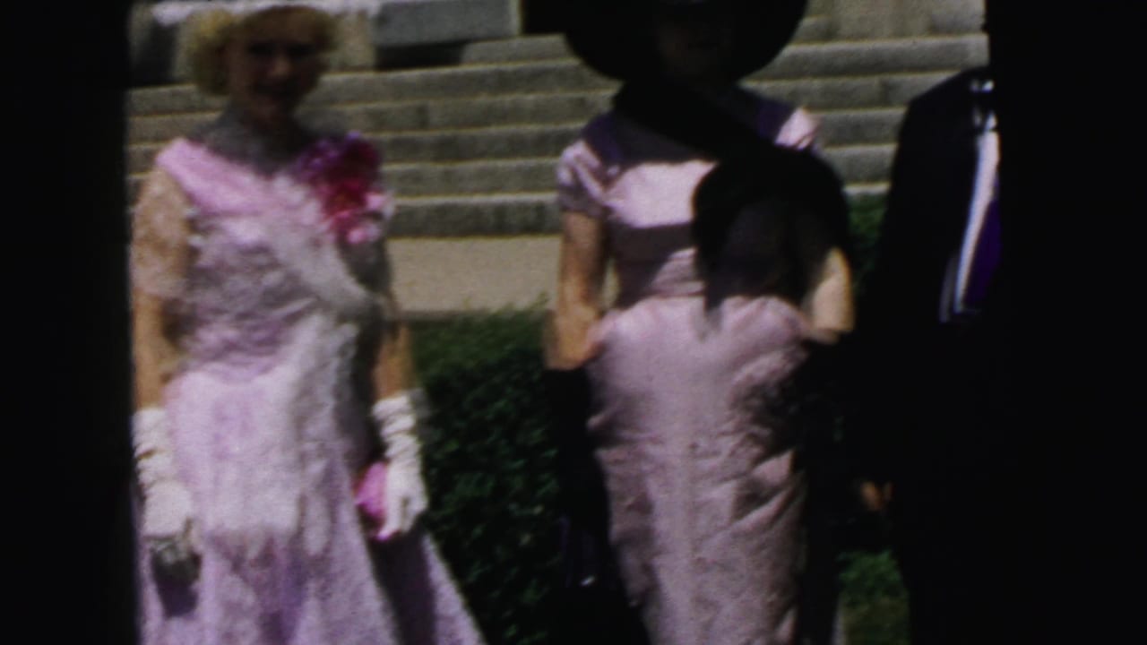 1964 Camden home-movie frame showing formally dressed women in pastel dresses and hats outside a building with stone steps.