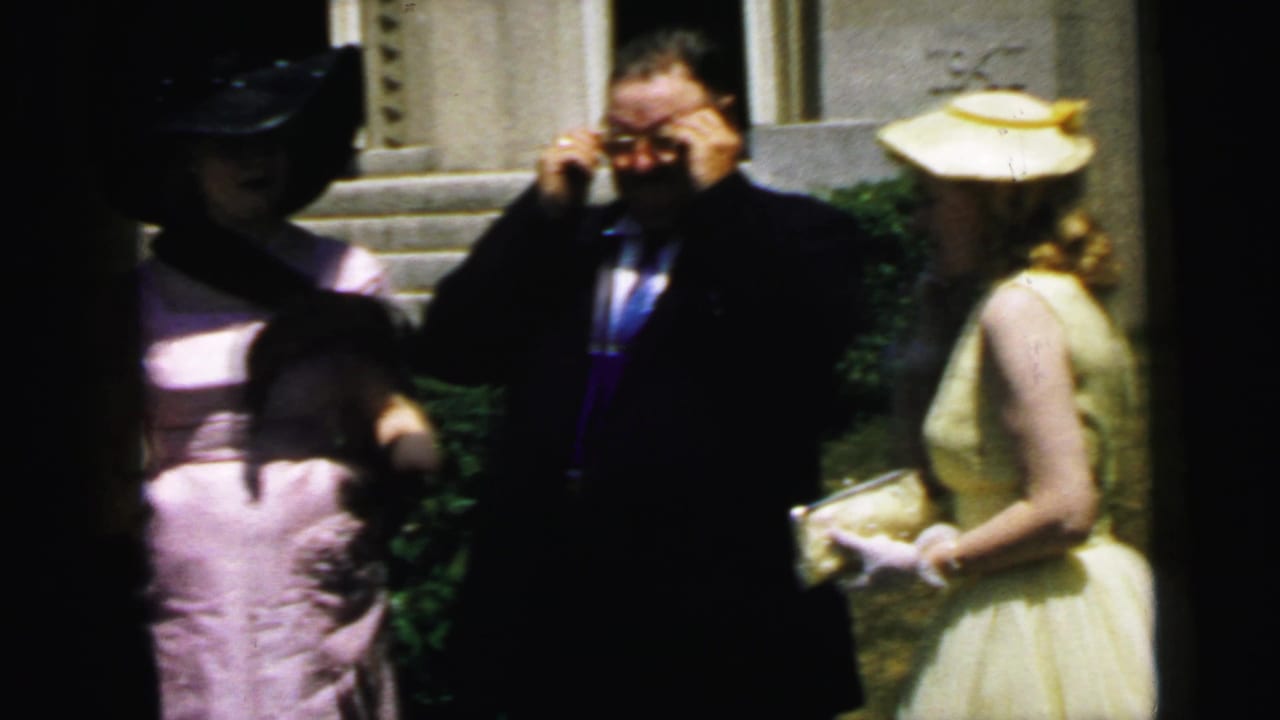 1964 Camden frame with a man in a dark suit adjusting his glasses near a woman in a pale yellow dress and matching hat.