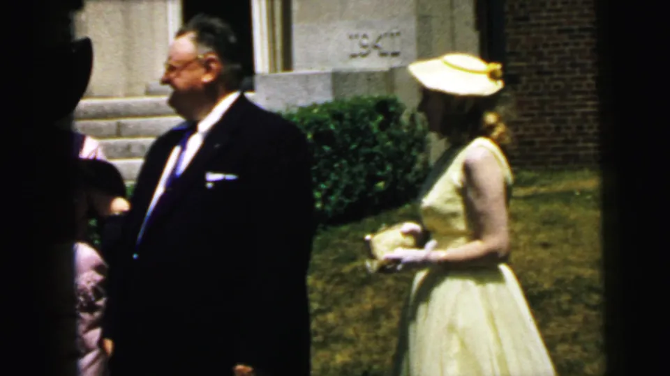 Home-movie frame from Camden in 1964 showing a suited man and a woman in a pale yellow formal dress near a building and lawn.