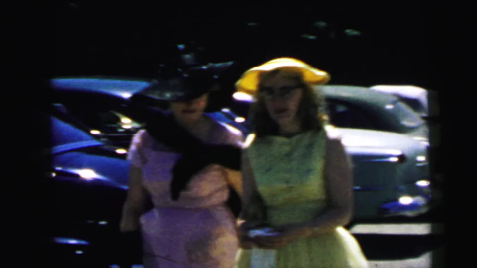 1964 Camden home-movie frame showing two formally dressed women in hats near parked mid-century cars, partly obscured by shadow.