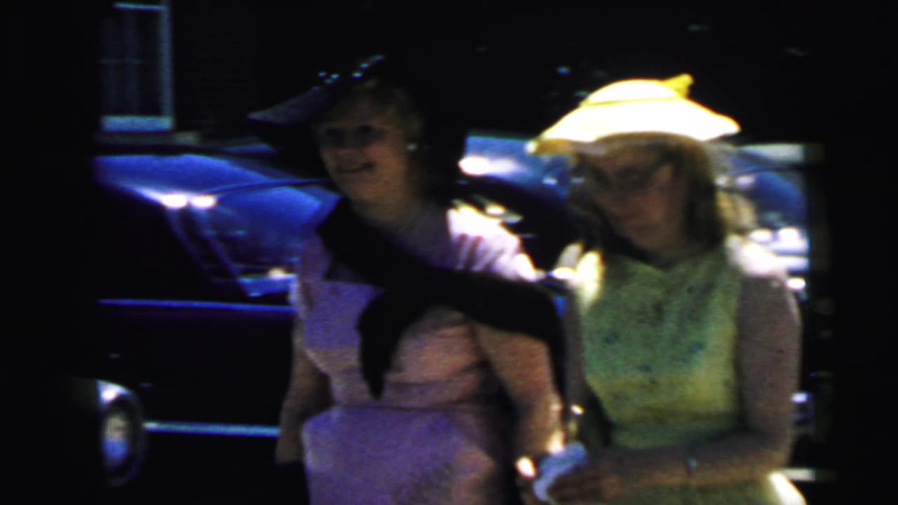 Two women in hats stand near parked cars while one holds a dark cat.