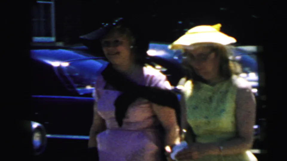 1964 Camden wedding home-movie frame showing two women in formal dresses and hats near parked cars, with one holding a dark cat.