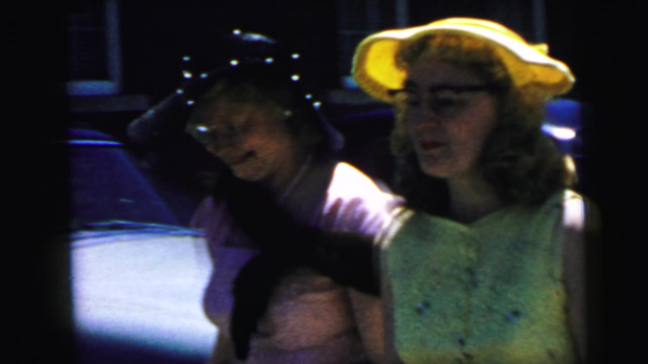 1964 Camden wedding footage shows two women in dress hats beside parked cars, with a dark cat stretched across one woman's arms.