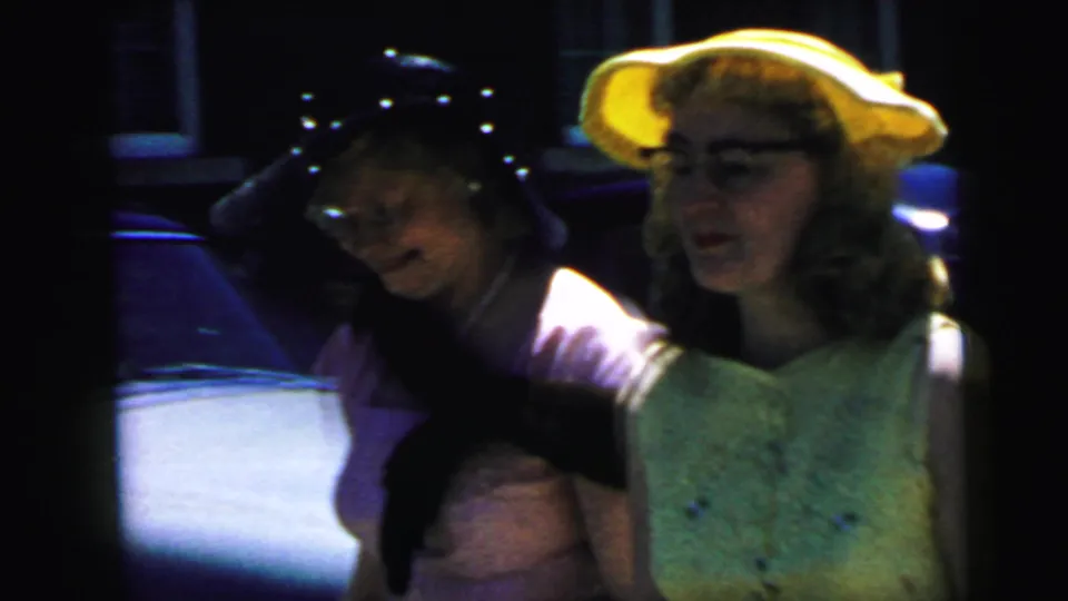 1964 Camden wedding footage shows two women in dress hats beside parked cars, with a dark cat stretched across one woman's arms.
