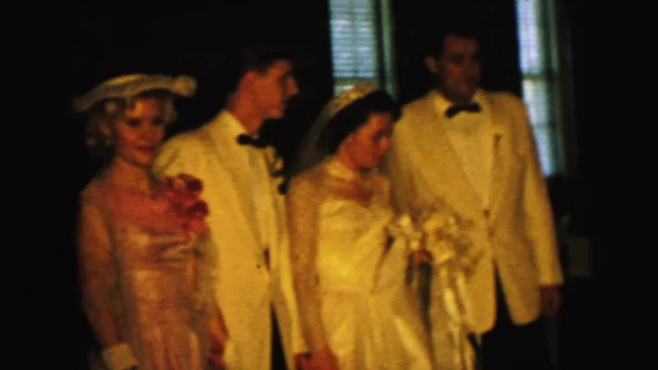 Indoor 1964 Camden wedding scene with bride in veil and gown, groom in a light jacket, and guests arranged for the home-movie camera.