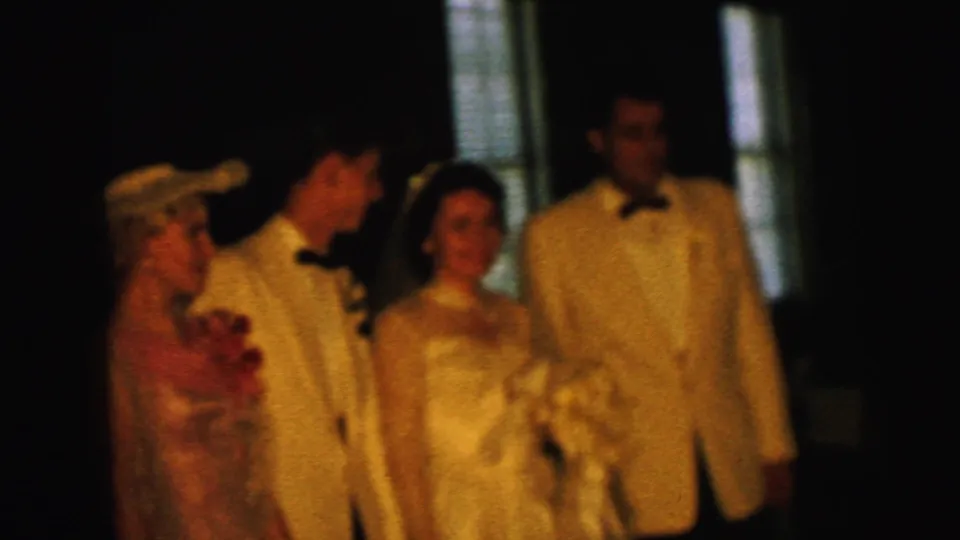 1964 Camden wedding home movie frame showing the bride and groom indoors with attendants in light jackets and a woman wearing a corsage.