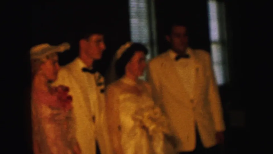 The bride, groom, bridesmaid, and tuxedoed man continue posing in a Camden 1964 wedding scene with dark interior surroundings.