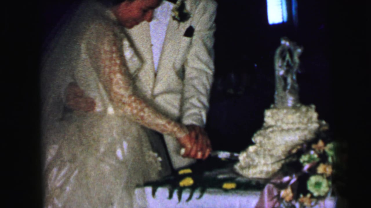 The newlyweds in the Camden, New Jersey home movie lean toward the wedding cake while guiding the knife together.