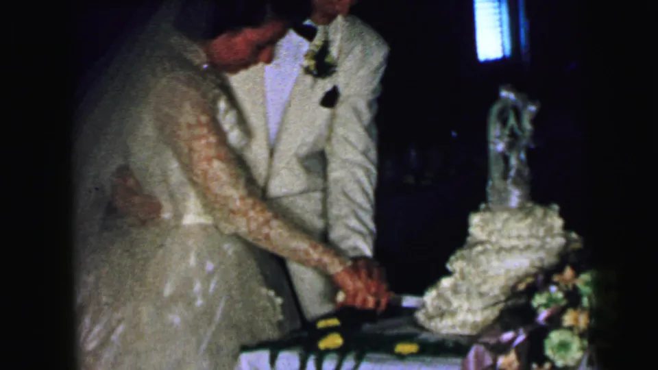 The bride looks down toward the cake knife while the groom stands close, their hands joined during the 1964 wedding cake ritual.