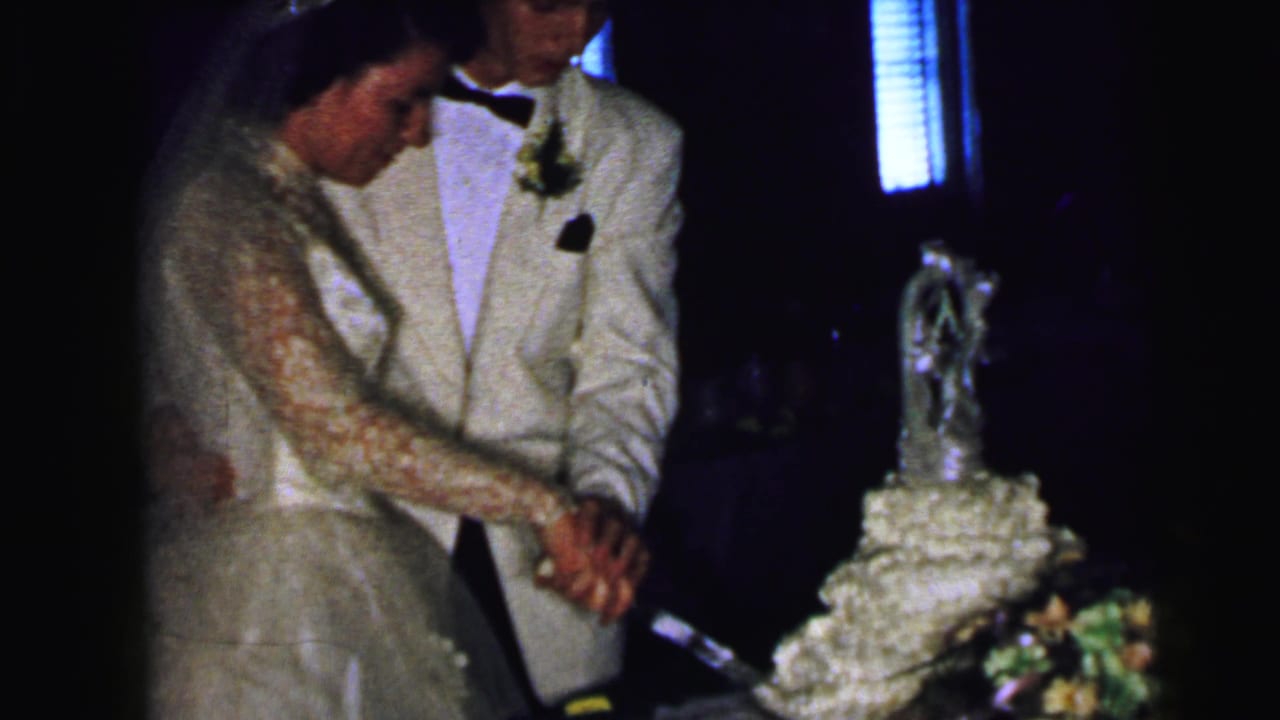 In this 1964 Camden, New Jersey 8mm frame, a bride in a lace gown and a groom in a light tuxedo begin cutting a tiered wedding cake.
