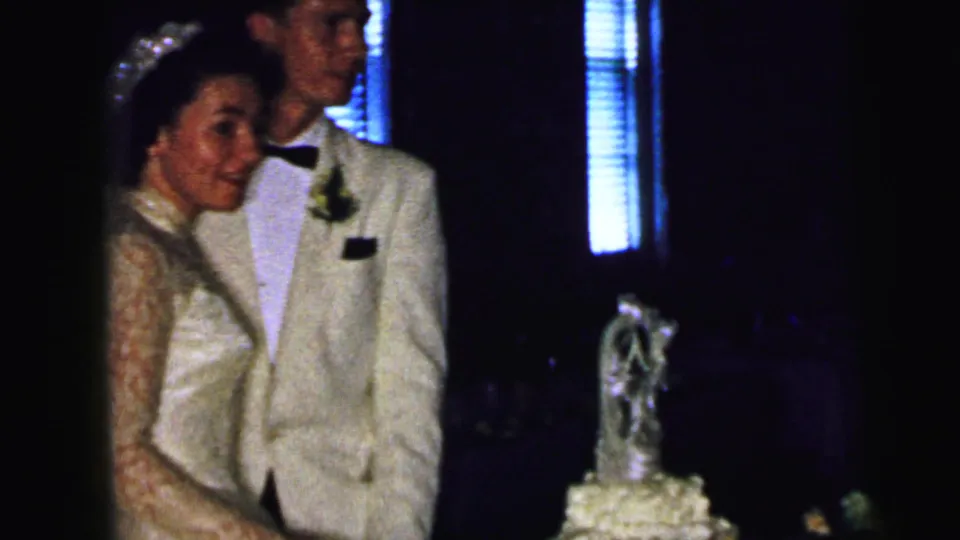 The bride smiles beside the groom and tiered cake, with dark interior lighting and bright blinds behind them.
