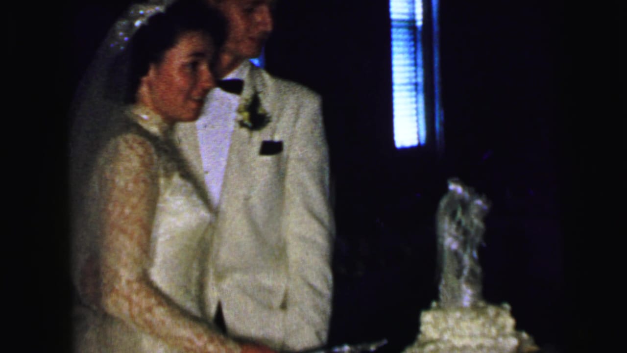 In supported 1964 Camden home-movie footage, a bride in a lace gown and groom in a light suit stand beside their wedding cake.