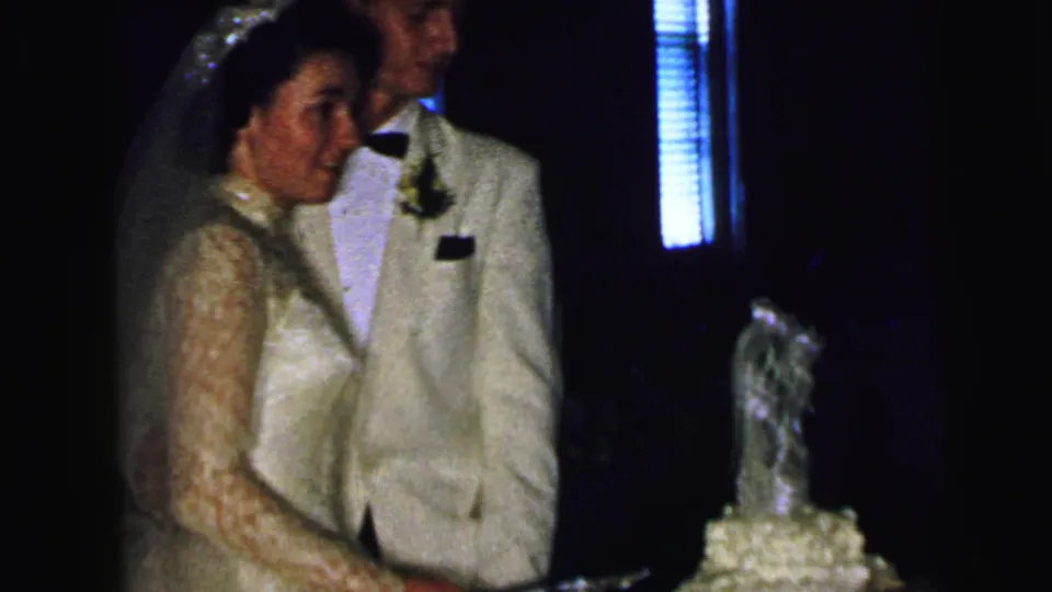 In supported 1964 Camden home-movie footage, a bride in a lace gown and groom in a light suit stand beside their wedding cake.