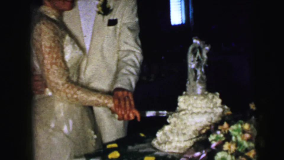 The bride and groom continue the cake-cutting gesture, framed close against a dark reception-room background in 1964 Camden footage.