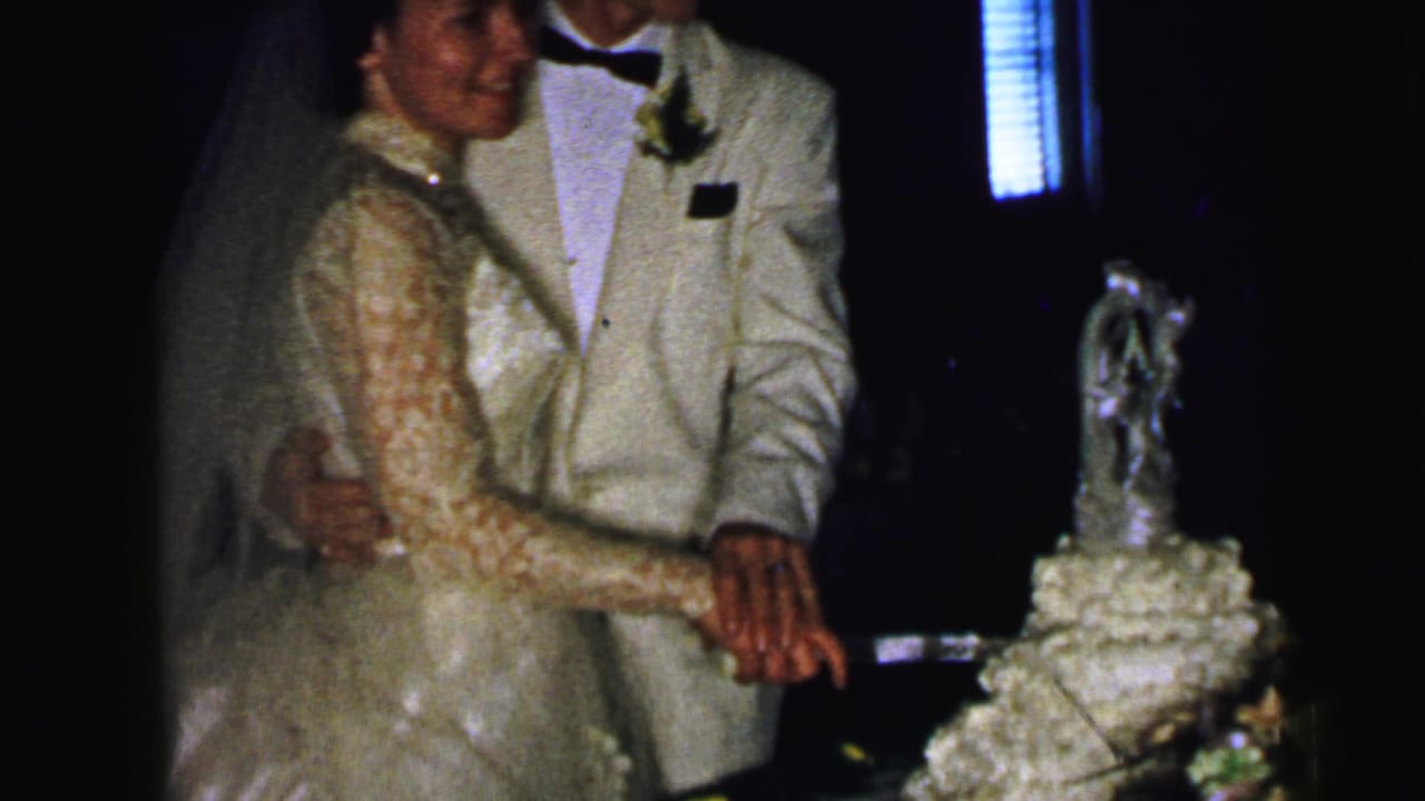 In 1964 Camden wedding reception footage, a bride in a lace gown and veil stands with her groom beside a tiered cake as they prepare to cut it.