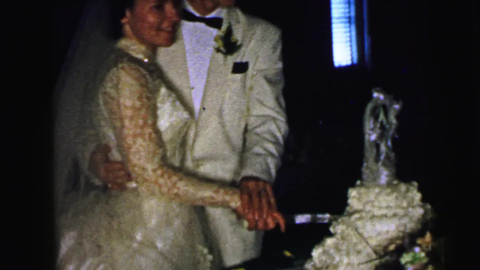 In 1964 Camden wedding reception footage, a bride in a lace gown and veil stands with her groom beside a tiered cake as they prepare to cut it.