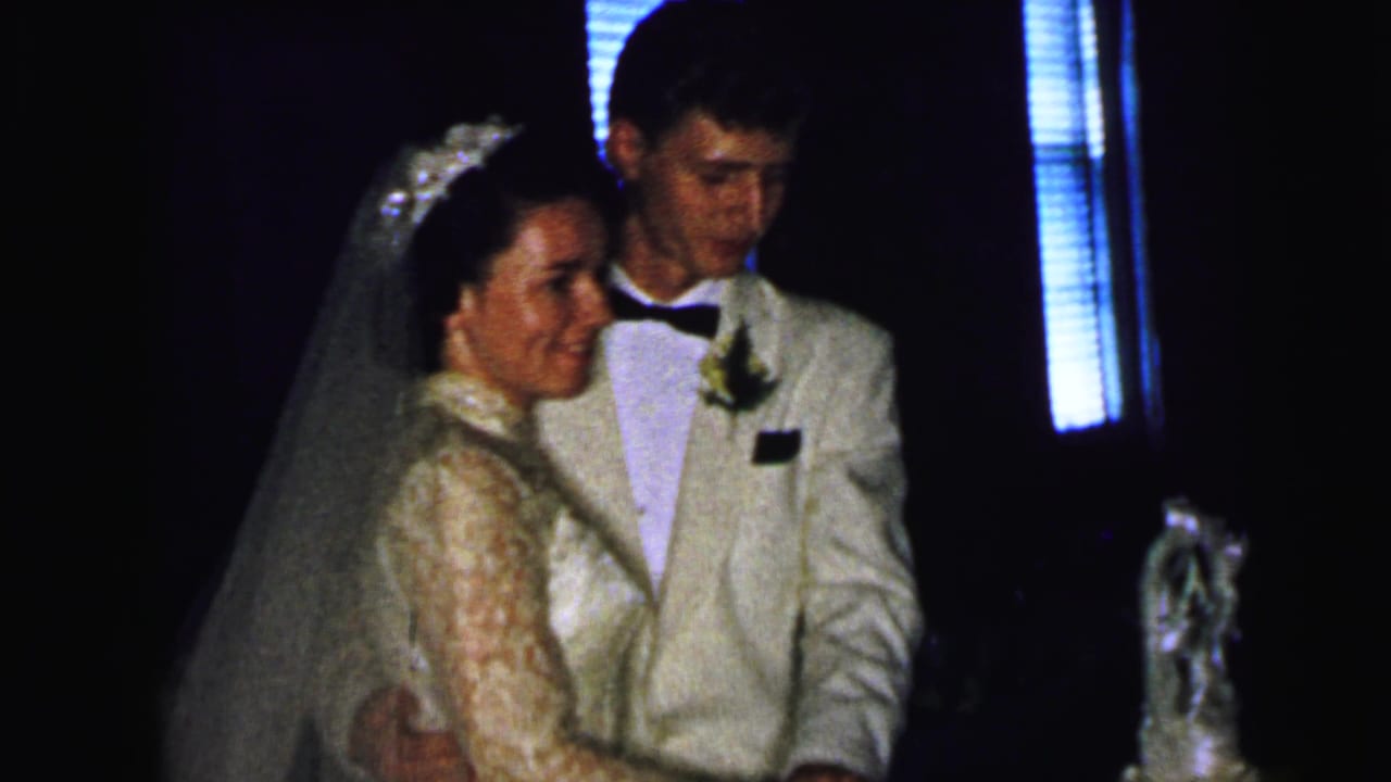 The bride and groom stand arm in arm near their wedding cake during a Camden, New Jersey reception recorded on 8mm film in 1964.
