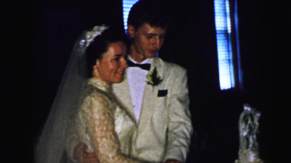 The bride and groom stand arm in arm near their wedding cake during a Camden, New Jersey reception recorded on 8mm film in 1964.