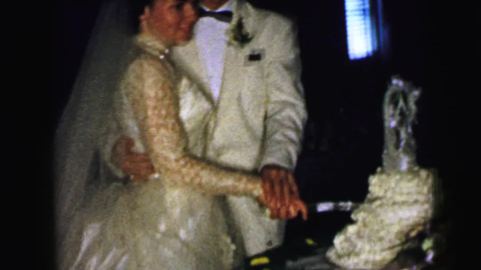 The couple jointly cuts the tiered wedding cake, with the bride in a lace gown and the groom in a white jacket visible in the reception interior.