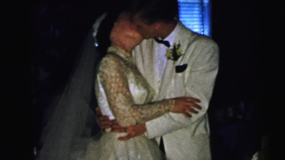 The bride and groom kiss while embracing beside the wedding cake in 1964 Camden reception footage, with the dark room and cake topper partly visible.