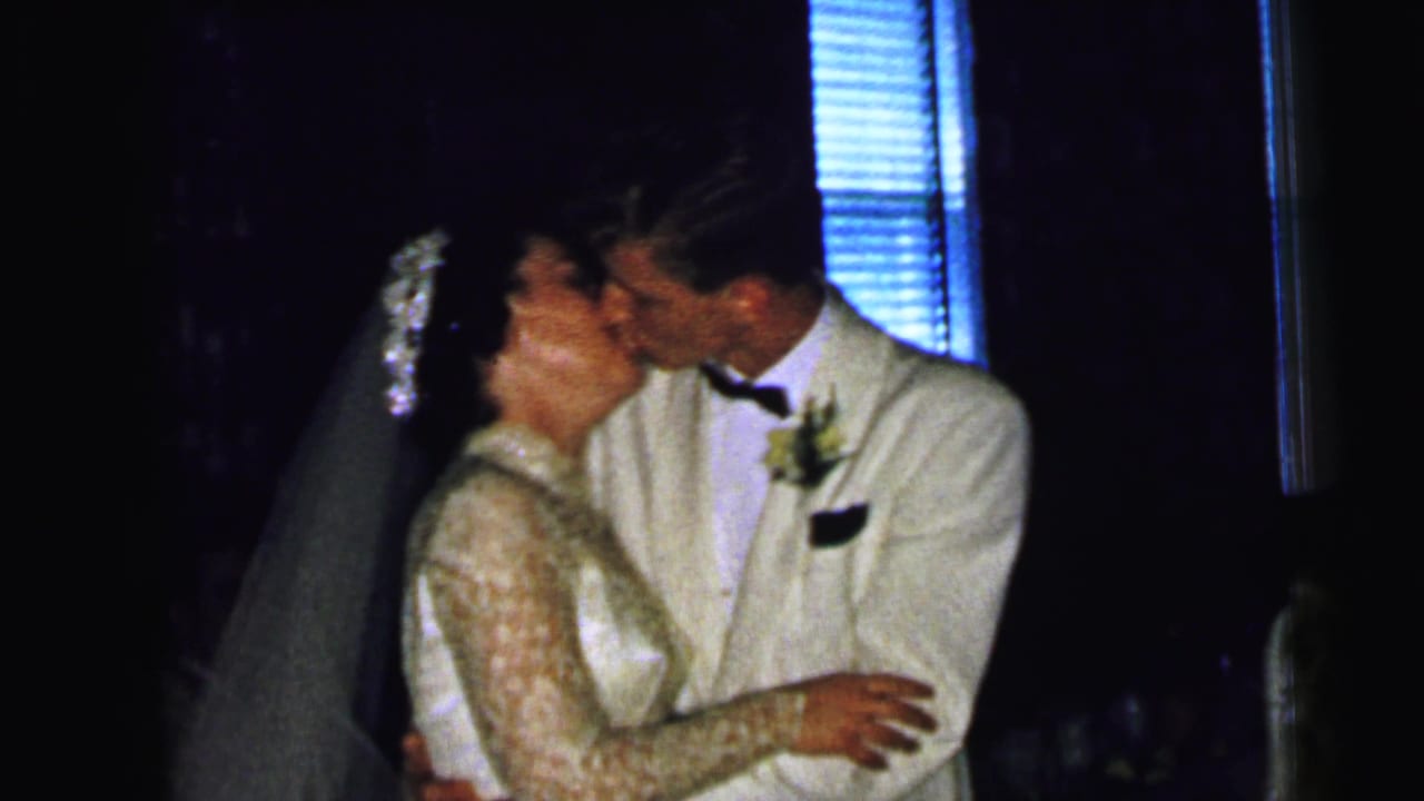 The bride and groom remain in a close embrace during the 1964 Camden wedding scene, with blinds visible behind them.