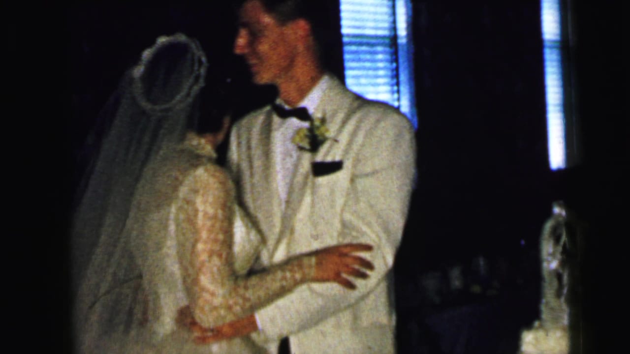 Bride and groom embrace while talking near window blinds.