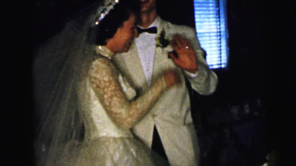 The bride gestures near the groom during a dim 1964 Camden wedding reception captured on 8mm film.